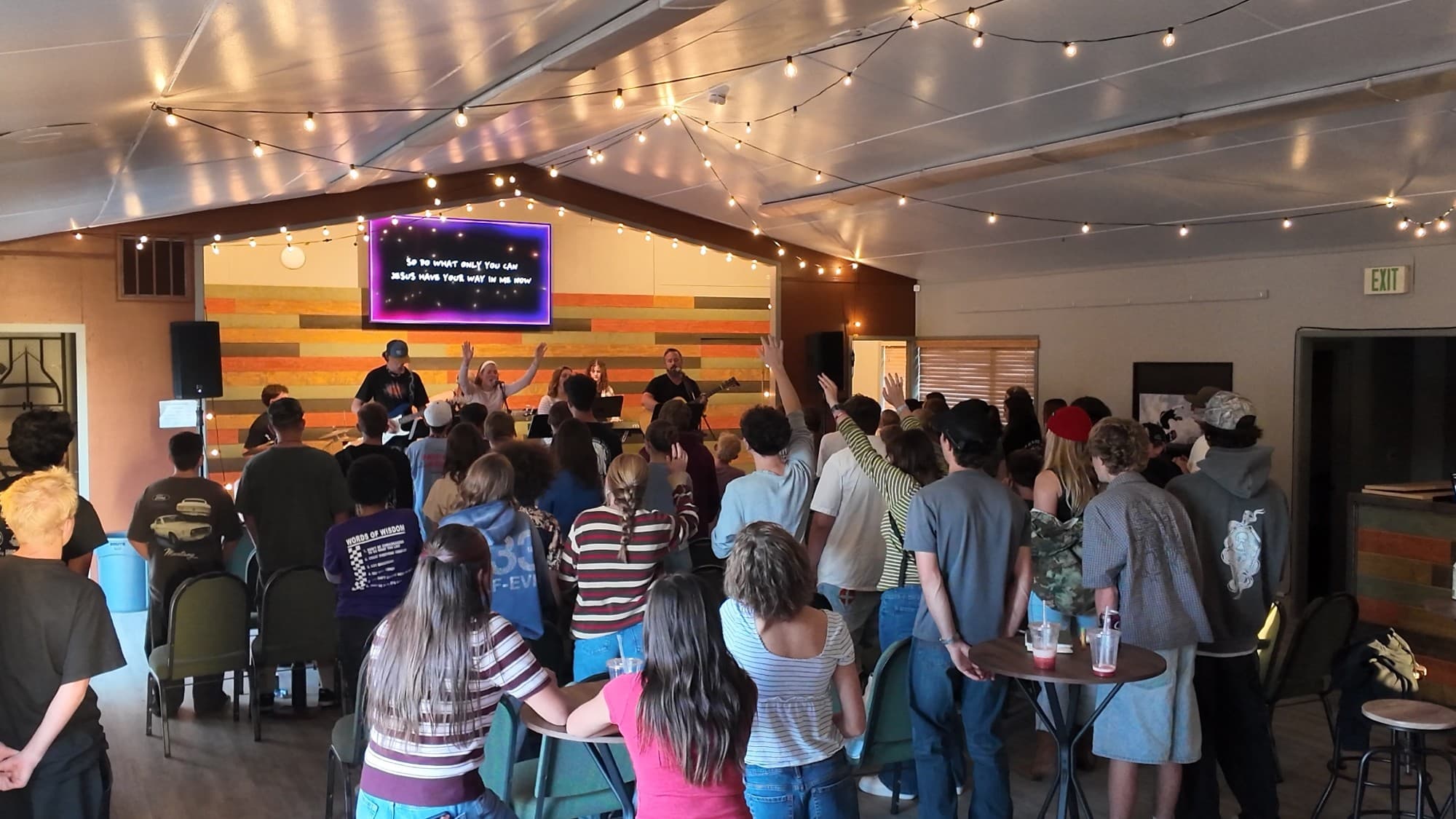 Students with hands raised in worship at Youth Night, with the band on stage under string lights